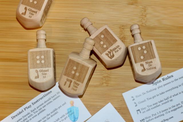 Wooden dreidel with braille letter matching the starting sound of the Hebrew letter on each side (N-Nun, H - Hey, G - Gimel, S - Shin) plus game instruction card and QR code link to audio game instructions.